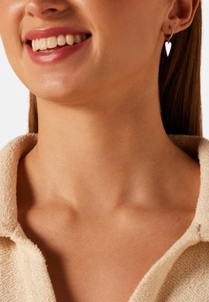 Smiling woman wearing a beige textured collar shirt and a silver hoop earring with a dangling heart charm on her right ear.