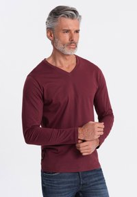 Camiseta de manga larga color marrón burdeos con escote en V, hecha de tela suave, combinada con jeans oscuros. Detalle sutil de etiqueta en el dobladillo lateral visible.