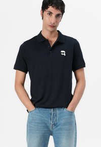 Mornarsko polo majica s kratkimi rokavi, s ovratnikom, tri gumbi na zapenjanju in belim logotipom na prsih. Denimske kavbojke dopolnjujejo videz.