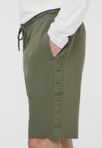 Pantaloni corti in cotone verde oliva con vita elasticizzata, cordino e tasca laterale. Il pannello laterale testurizzato presenta la scritta debossata "EVERYONE."