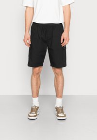 Černé šortky s elastickým pasem, s bočními kapsami a nenápadným brandingem. Nosené s bílým tričkem a hnědými teniskami.