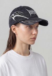 Donna che indossa un cappellino da baseball nero consumato con disegni grafici bianchi e scritte, abbinato a una maglietta bianca girocollo semplice.