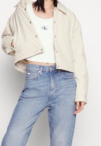 Hellbeige gekürzte Jacke mit Druckknöpfen und klassischem Kragen, kombiniert mit einem weißen Tanktop und hellblauen, hoch taillierten Jeans.
