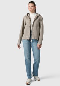 Giacca con cappuccio grigia chiara, dotata di zip, due tasche frontali e caratteristiche regolabili, abbinata a jeans blu e sneakers bianche.