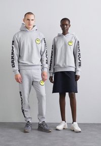 Grå hettegensere med store smiley-lapper, svart tekst, kombinert med grå joggebukser og et mørkt skjørt. Fotwear inkluderer grå og hvite joggesko.