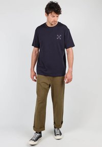 T-shirt bleu marine à manches courtes avec un petit motif brodé sur la poitrine, associé à un pantalon vert olive et des baskets noires.
