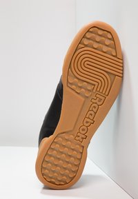 Svart sneaker med en teksturert gummisåle, med et geometrisk mønster og preget "Reebok"-logo. Gummimateriale og slank design.