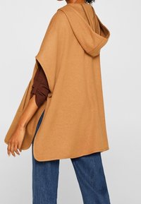 Mujer con una capa holgada color camello con capucha sobre una camiseta de manga larga marrón y vaqueros azules, vista desde atrás.