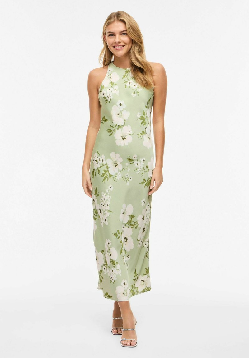 Mujer rubia sonriendo, vestida con un maxi vestido sin mangas de color verde claro con estampado floral y sandalias plateadas de tacón con tiras, posando frente a un fondo blanco.