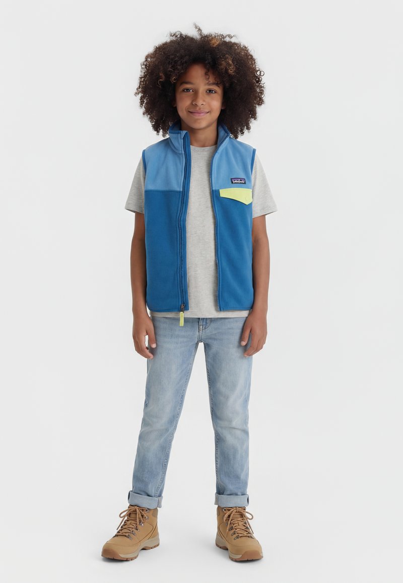 Παιδί με κατσαρά μαλλιά φοράει γαλάζιο φλις γιλέκο, γκρι μπλουζάκι, ανοιχτόχρωμο τζιν και καφέ μπότες, στέκεται μπροστά με άσπρο φόντο.