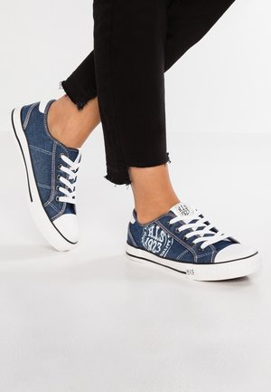 Osoba nosząca niebieskie, niskie trampki z dżinsu z białymi sznurowadłami i podeszwami, stojąca na białej podłodze, ubrana w czarne, krótkie spodnie.