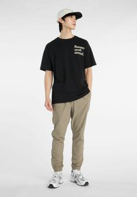 Camiseta negra de manga corta con el texto "Los corredores no son normales". Pantalones jogger beiges con puños elásticos. Zapatillas deportivas blancas. Gorra clara.