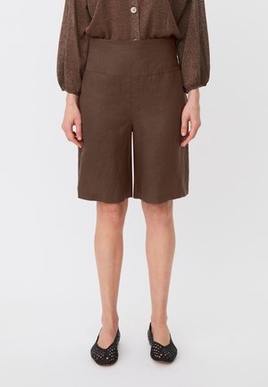 Mujer con pantalones cortos marrones de talle alto hasta la rodilla, con una blusa marrón de manga larga y suelta y zapatos planos negros tejidos, de pie sobre un fondo blanco.