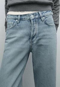 Vaaleansiniset farkut rentoa istuvuutta, joissa on nappisulku, viisi taskua ja hienovarainen haalistuma reisien kohdalla. Kankaan tekstuurinen yksityiskohta näkyvissä.