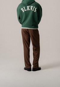 Persona con sudadera verde con el texto "BLKVIS", pantalones marrones y zapatos negros, de pie contra un fondo claro y liso, de espaldas.