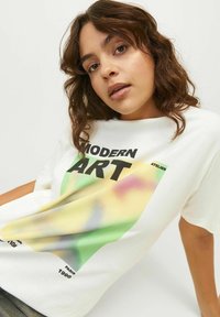 Camiseta de algodón blanca de manga corta que presenta un gran estampado gráfico en verde, amarillo y rosa, con las palabras "ARTICULO MODERNO" en letras negras en negrita.