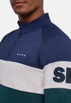 Mann trägt ein farblich abgesetztes Viertel-Reißverschluss-Sweatshirt in Marineblau, Weiß und Grün mit dem Schriftzug "FALKE" auf der Brust und teilweisem "SI"-Schriftzug am Ärmel.
