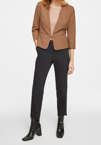 Femme portant un blazer marron, un haut à motifs, un pantalon noir jusqu'aux chevilles et des bottines noires à talons, debout les mains dans les poches.