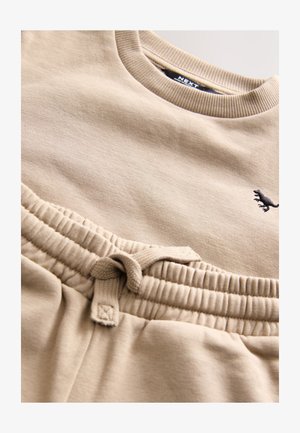 Conjunto de sudadera y pantalones deportivos beige en mezcla suave de algodón. Cuenta con cuello acanalado, cintura elástica con cordón y pequeño detalle de logo.