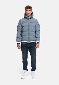 Light blue puffer jacket with a zip front, two chest pockets, ribbed cuffs and hem, and a hood. Worn with dark jeans and white sneakers.