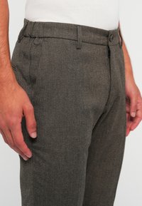 Prim-plan al unei persoane îmbrăcate cu pantaloni gri texturat, cu buzunare frontale și talie cu nasture, asortați cu o cămașă albă.
