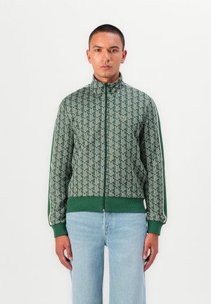 Jachetă verde cu fermoar, prezentând un model geometric, manșete și tiv ribbate, dungi albe pe laterale și un logo mic pe pieptul stâng.
