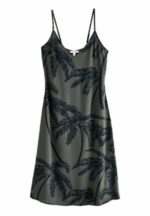 Vrouw draagt een mouwloze groene jurk met zwarte palmboom motieven, staand bij een witte bank met beige kussens in een zonnige kamer.