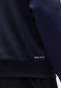 Κοντινή λήψη σε ύφασμα αθλητικής ένδυσης σε ναυτικό μπλε με το λευκό λογότυπο "DRI-FIT" κεντημένο κοντά στο τελείωμα πάνω από σκούρο γκρι παντελόνι.