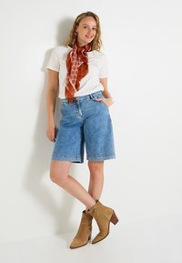 Camiseta blanca con logo, bufanda marrón con estampado, pantalones cortos de mezclilla azul claro y botas de ante color tostado con tacón bloque.