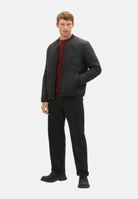 Schwarze gesteppte Bomberjacke mit Reißverschluss, gerippten Bündchen und rundem Kragen, getragen über einem burgunderfarbenen Pullover und kombiniert mit schwarzen Hosen.