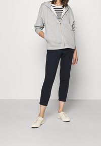 Felpa grigia con zip sopra una maglietta a righe bianco e nera, abbinata a pantaloni blu corti e sneaker color crema chiaro. Design semplice e casual.