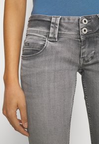 Jeans de mezclilla gris con un diseño elegante, que cuentan con dos cierres de botón, una pequeña etiqueta con logo en la cintura y un bolsillo frontal.