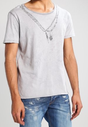 Grå, kortærmet t-shirt lavet af blødt stof, med et kædegrafikdesign med et vedhæng, parret med slidte blå denimjeans.