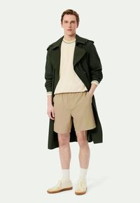 Tume roheline trench coat, kreemjasvest, tan värvi lühikesed püksid, valged sokid, kollased tossud. Mudel seisab sirgelt, käed taskutes, neutraalne taust.