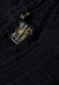 Tissu bleu marine avec un design texturé et froissé. Présente une étiquette Superdry en évidence avec un graphique de soleil et de palmier en broderie dorée et argentée.