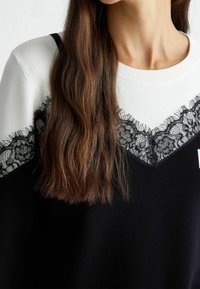 Pull noir et blanc avec un col en V, orné d'un détail en dentelle le long du col. Confectionné dans un tissu doux et texturé.