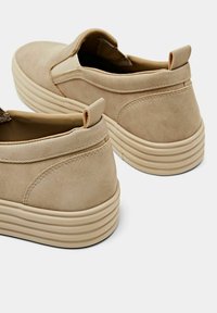 Paire de chaussures décontractées beige à enfiler avec semelles épaisses rainurées et languettes aux talons, vues de trois quarts arrière.