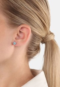 Kvinna med blont hår i hästsvans som bär en rund aquamarinörring i vänster öra mot en vit bakgrund.