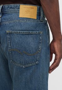 Πίσω τσέπη και ετικέτα σε μπλε τζιν Jack & Jones που φοράει ένα άτομο με σκουρόχρωμο δέρμα και σκουρόχρωμο πουκάμισο.