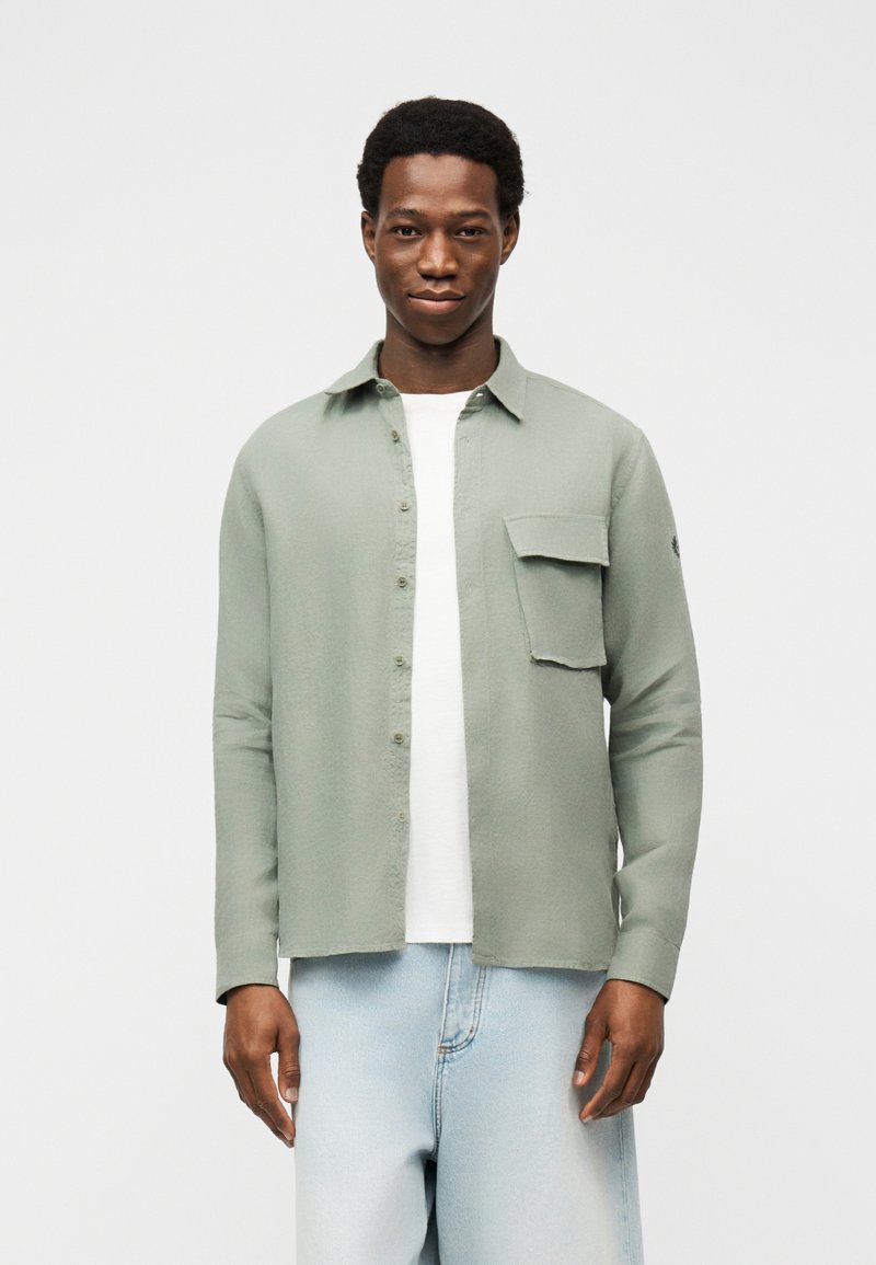 Άνδρας που φοράει ανοιχτό πράσινο πουκάμισο με κουμπιά πάνω από λευκό μπλουζάκι και ανοιχτό μπλε τζιν, στέκεται μπροστά σε απλό λευκό φόντο.