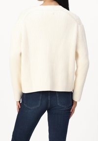 Cremiger, gerippter Pullover mit langen Ärmeln und lockerer Passform, ausgestattet mit einem Rundhalsausschnitt und einem leicht verkürzten Saum, kombiniert mit dunklen Jeans.