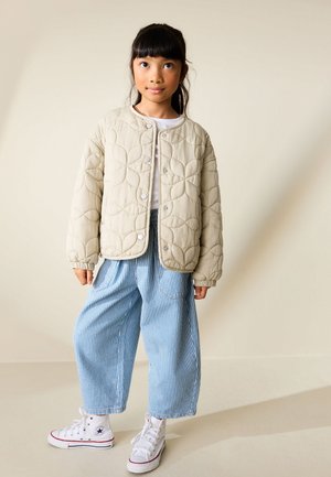 Joven chica con cabello negro que lleva una chaqueta acolchada beige, camiseta blanca, pantalones anchos a rayas azules y zapatillas blancas de caña alta, de pie en el interior.