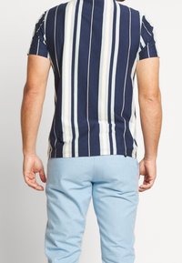 Muž oblečený v košili s krátkým rukávem modro-bílé vertikální pruhy a světle modrých kalhotách, zobrazený zezadu na bílém pozadí.