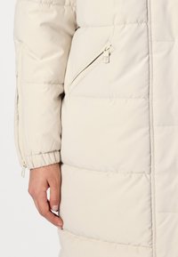 Manteau matelassé beige clair présentant une texture douce et quiltée, une poche zippée inclinée et des poignets élastiqués pour un ajustement sécurisé.