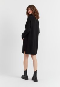 Schwarz gestricktes Kleid mit hohem Hals, langen Ärmeln, seitlichen Schlitzen und roten Akzenten am Saum. Trägt man mit schwarzen Stiefeletten. Glatte Textur.
