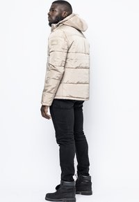 Giubbotto imbottito beige con cappuccio, design a righe orizzontali e un'etichetta sulla parte superiore del braccio. Indossato con jeans neri e stivali.