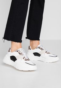 Zapatillas blancas con acentos de estampado de leopardo, paneles en negro y gris, suelas texturizadas y cordones redondos, exhibidas con unos vaqueros negros recortados.