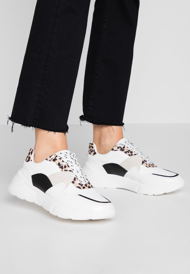 Zapatillas blancas con acentos de estampado de leopardo, paneles en negro y gris, suelas texturizadas y cordones redondos, exhibidas con unos vaqueros negros recortados.