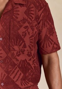 Chemise rouge à manches courtes en tissu texturé présentant un motif répétitif floral et géométrique, boutons sur le devant et poche plaquée.