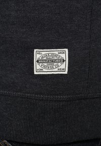 Etichetta in tessuto grigio scuro con il testo "JACK & JONES" delineato in bianco, su uno sfondo testurizzato con dettagli di cucitura.